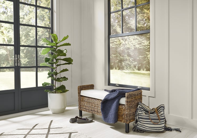 Sunlit entryway with white paneled walls, black-framed windows, a wicker bench, and a potted fiddle-leaf fig